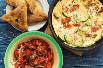 Fried omelette with zucchini, leeks and tomatoes served in a frying pan with marinated tomatoes with onions and pies on a rustic wooden background.
