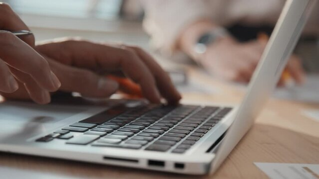 Close up slowmo side shot of unrecognizable male hands entering data into laptop and using keyboard and touchpad in light office
