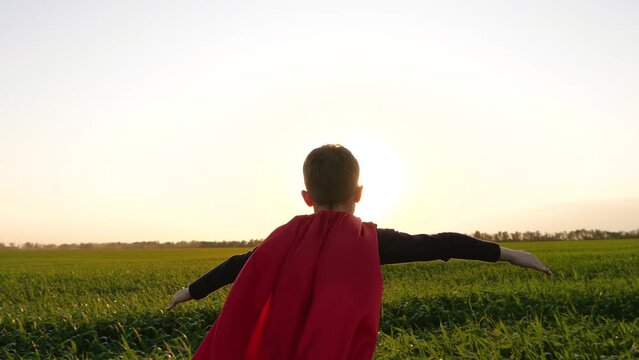kid superhero running through a green grass. Boy in red cape view from back running in sunset sunlight. kids dream concept, freedom, outstretched