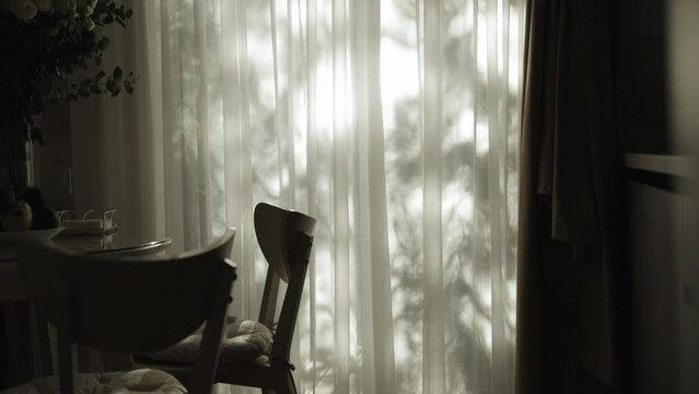 Close-up silhouette of kitchen interior with wooden round table, vase with flowers on it, four wooden chairs near the large window and sun rays through the tulle on the chairs.