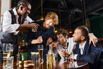 happy interracial friends laughing and drinking alcohol drinks while relaxing after work in bar