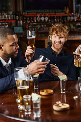 cheerful redhead man pointing at friend while having drinks after work in bar, interracial friends