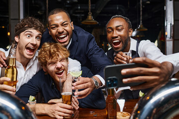 Fototapeta na wymiar excited multiethnic colleagues in formal wear taking selfie on smartphone in bar after work