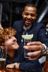 focus on tequila shot with sliced lime on top, african american man holding drink near friend in bar