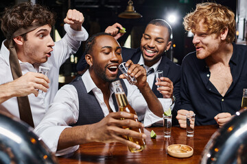 happy interracial men cheering near african american friend drinking tequila shot and holding beer