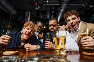 emotional multicultural colleagues in formal wear holding beer and screaming in bar, men after work