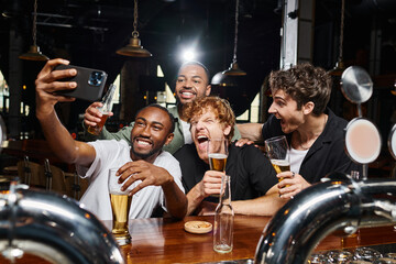 happy multicultural men taking selfie on smartphone while drinking beer in bar, bachelor party