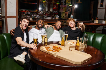 four cheerful interracial friends hugging and sitting near pizza and beer, spending time in bar