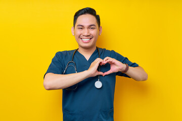 Smiling professional young Asian man doctor or nurse wearing a blue uniform and stethoscope showing heart shape with hands isolated on yellow background. Healthcare medicine concept