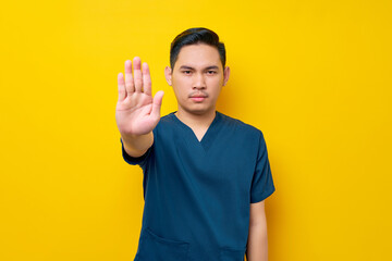 Serious professional young Asian male doctor or nurse wearing a blue uniform makes a stop gesture with his hands isolated on yellow background. Healthcare medicine concept - obrazy, fototapety, plakaty