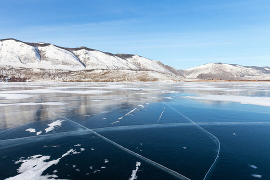 Sunny winter day on frozen Baikal Lake. Small Sea is covered with beautiful clear smooth blue ice with cracks. In distance snow-covered mountains and Sarma village. Beautiful winter landscape