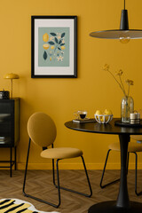 Stylish composition of warm dinning room interior with mock up poster frame, round table, yellow chair, glass vase with dried flowers, lamp, and personal accessories. Home decor. Template.