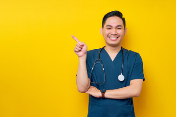 Smiling professional young Asian male doctor or nurse wearing a blue uniform standing confident while pointing a finger at copy space isolated on yellow background. Healthcare medicine concept