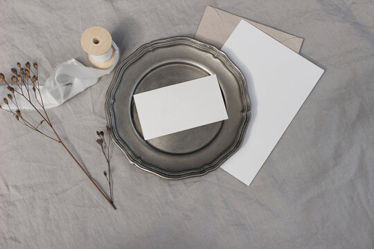 Vintage neutral wedding stationery. Blank Christmas greeting card, invitation mockup on old silver plate. Dry flower, craft paper envelope in sunlight. Linen tablecloth, silk ribbon. Winter