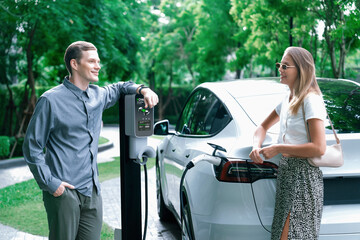 Young couple travel with EV electric car charging in green sustainable city outdoor garden in...