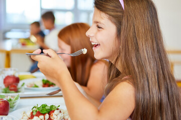 Happy girl eating meal with fork in school cafeteria