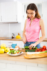 Positive woman is cutting vegetables for salad in kitchen at home
