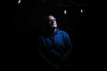 Senior man wearing blue sweater picked out by spotlight in dark room