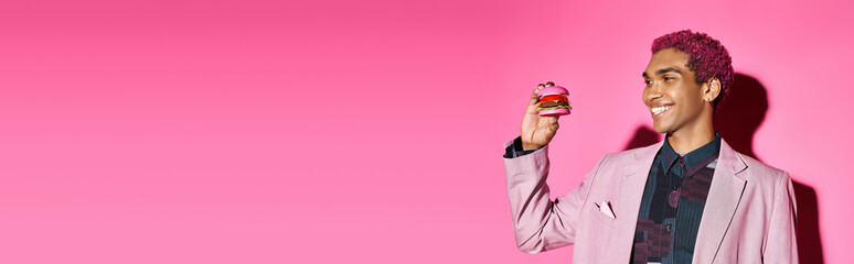 joyful handsome man with curly hair looking cheerfully at mini burger on pink background, banner