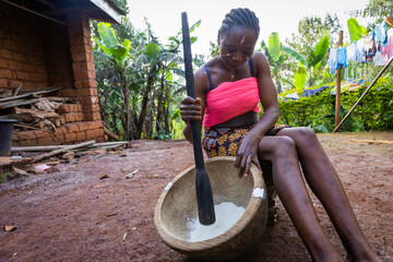 Young African woman living in a rural area pounding yam to cook fufu for dinner - obrazy, fototapety, plakaty
