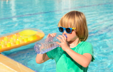 Cute little boy drinks water from a plastic bottle against the background of a swimming pool....