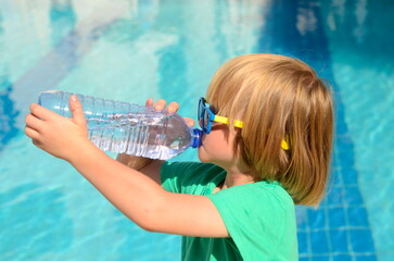 Cute little boy drinks water from a plastic bottle against the background of a swimming pool....