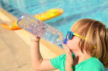 Cute little boy drinks water from a plastic bottle against the background of a swimming pool....