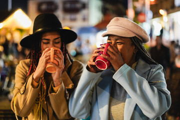 African mother and daughter having drinking hot chocolate outdoor at winter market - Focus on senior female hand holding cup - obrazy, fototapety, plakaty