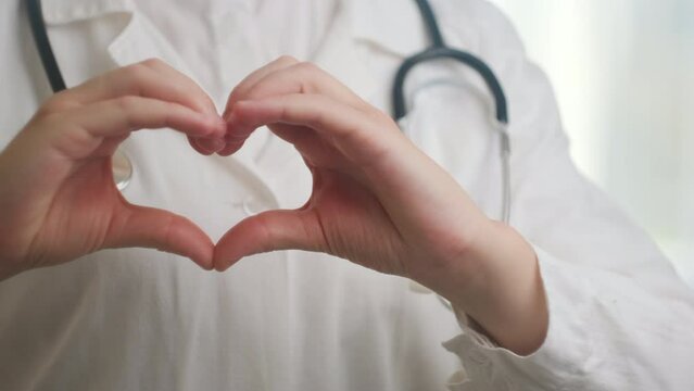 Close up of unknown woman doctor cardiologist in uniform with stethoscope showing heart symbol with fingers, expressing love and support to patients, healthcare medical help charity donation concept