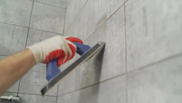 Tilers fill the space between the tiles with a rubber putty knife. Close-up of a hand in a protective glove holding a yellow trowel while grouting tiles. Grouting joints in the bathroom. Grouting