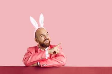 Funny man in suit and headband with Easter bunny ears shows on copy space on pink background. Caucasian young bald bearded man in pink formal suit points finger to side. Advertising banner. - obrazy, fototapety, plakaty