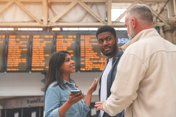 Group of multiracial friends checking time on departure board at a railway train station before boarding train. Enjoying travel by train concept - obrazy, fototapety, plakaty