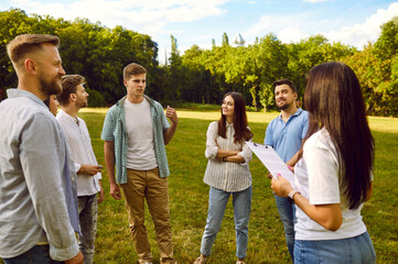 Brunette woman taking interview from a group of a young confident people students standing in a circle on green grass in the summer park and answering on social survey or having a training outdoors.