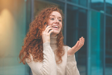 Portrait of beautiful happy young woman graduate with curly red hair talking to a friend on the...