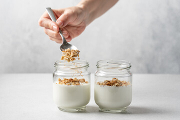 Female hands pouring granola into glasses with yogurt. Cooking breakfast, dessert parfait - obrazy, fototapety, plakaty