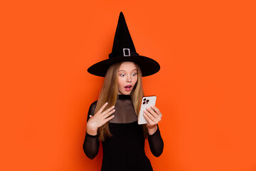 Photo portrait of lovely blonde teen lady amazed reaction hold gadget dressed black halloween garment isolated on orange color background