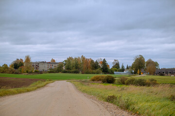 Latvia autumn landscape with sand and gravel road, agricultural field and small village between the trees