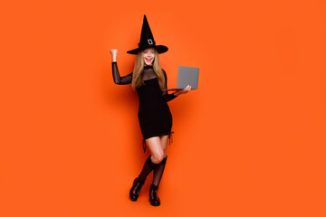 Full body photo of attractive blonde teen woman raise fist netbook dressed black halloween clothes isolated on orange color background