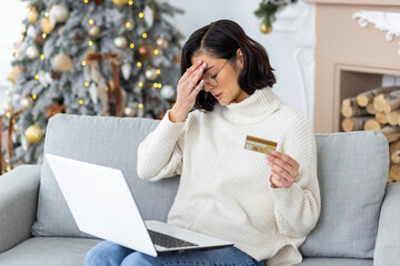 Sad young woman sitting at home on the sofa for the holidays near the Christmas tree, holding a...