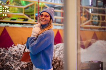 Cheerful and stylish woman, dressed in warm clothes, is having fun in a snowy winter amusement park