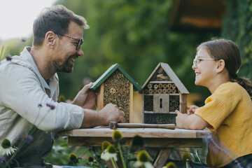 Father with daughter making bug hotel, or insect house outdoors in the garden. Girl learning about insects, garden ecosystem and biodiversity. - obrazy, fototapety, plakaty