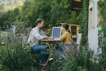 Father with daughter sitting outdoors in garden, with model of solar panel. Solar energy and...