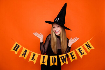 Photo of impressed funny girl dressed dark witch dress headwear hanging halloween decorations isolated orange color background
