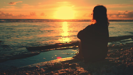 Woman sitting on beach looking at sunset sky and ocean waves. Dreaming girl enjoying summer vacation on a tropical island. Female silhouette, bright colorful sea view, travel, vacation