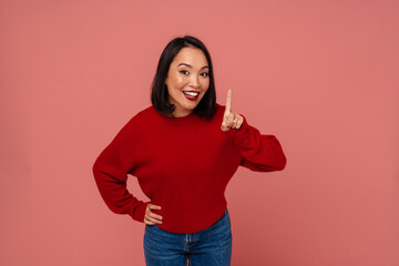 Smiling asian woman pointing finger up expressing she has idea while standing isolated over pink wall