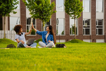 Two female students giving high five to each other while sitting on lawn in campus