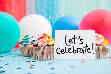 Let’s celebrate - lettering on blue background with colorful decorations, rainbow cupcakes and air balloons 