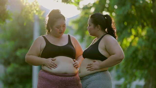 Overweight asian woman stressing over fat belly uses her hand to squeeze the excess fat while workout exercise in the morning. Sun rays shine through trees. 