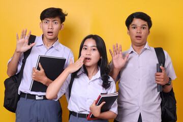 Shocked young Asian high school students holding books and carrying backpacks isolated on yellow...