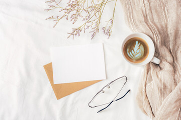Coffee cup, envelope with blank card, glasses and small flowers on white linen. Cozy workplace, breakfast, comfort, hygge concept. Top view, flat lay, mockup